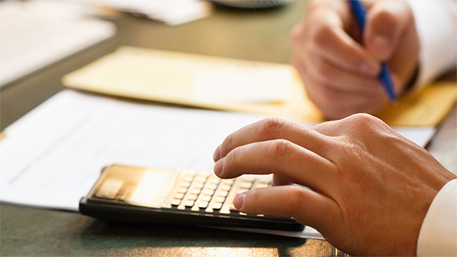 Person am Schreibtisch mit Taschenrechner und Stift (verweist auf: Kosten-Nutzen-Vergleich)
