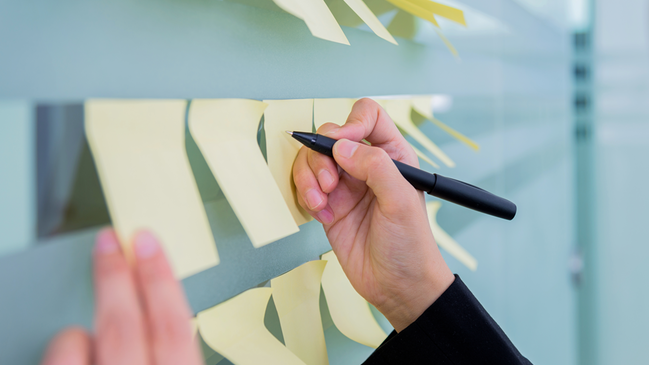 verweist auf: Aufgaben Eine Hand mit Stift schreibt etwas auf eine Pinnwand (verweist auf: Aufgaben)