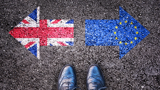 Eine Person steht vor zwei auf den Asphalt gemalten, gegenläufigen Pfeilen, die die britische und die EU-Flagge symobilisieren. (verweist auf: Brexit)