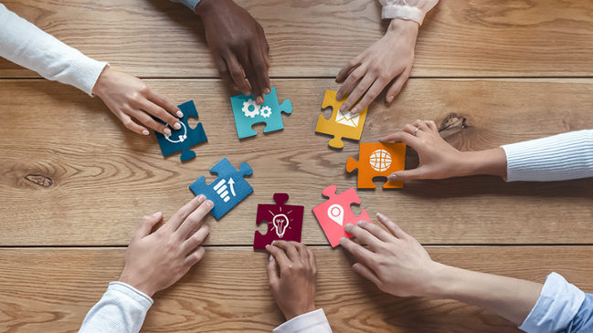 7 Hände die greifen nach bunten Puzzelstücken die auf einem Tisch liegen. 7 Hände die greifen nach bunten Puzzelstücken die auf einem Tisch liegen.